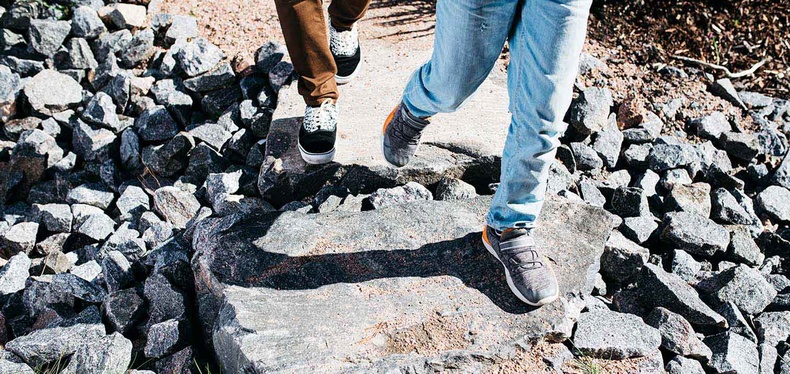 Kahden henkilön jalat kuvattuna yläviistosta. Maassa kiviä.